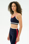 A woman with curly hair wears the SPLITS59 Eva Rigor Double Strap Bra in Indigo/White with matching leggings, both featuring contrast color details, against a plain white background.