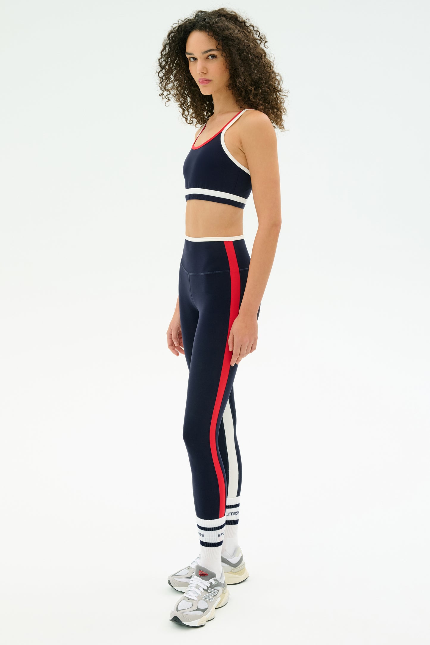 A woman models the SPLITS59 Eva Rigor Double Strap Bra in Indigo/White with white sneakers and curly hair, standing against a plain white background—showcasing sporty comfort and confidence for workouts or daily wear.