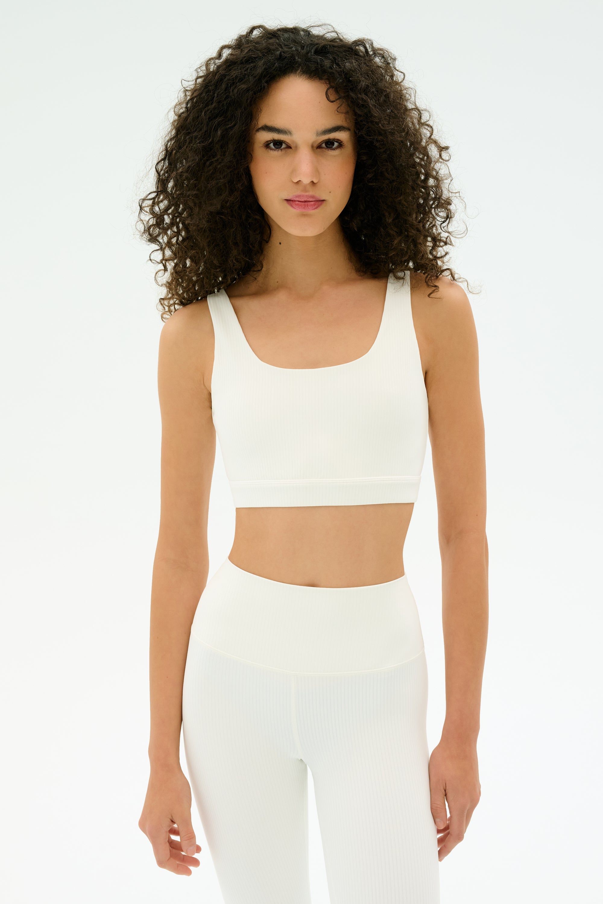 A woman with curly hair wears the SPLITS59 Airweight Rib Bra and matching high-waisted leggings, standing against a plain white background.