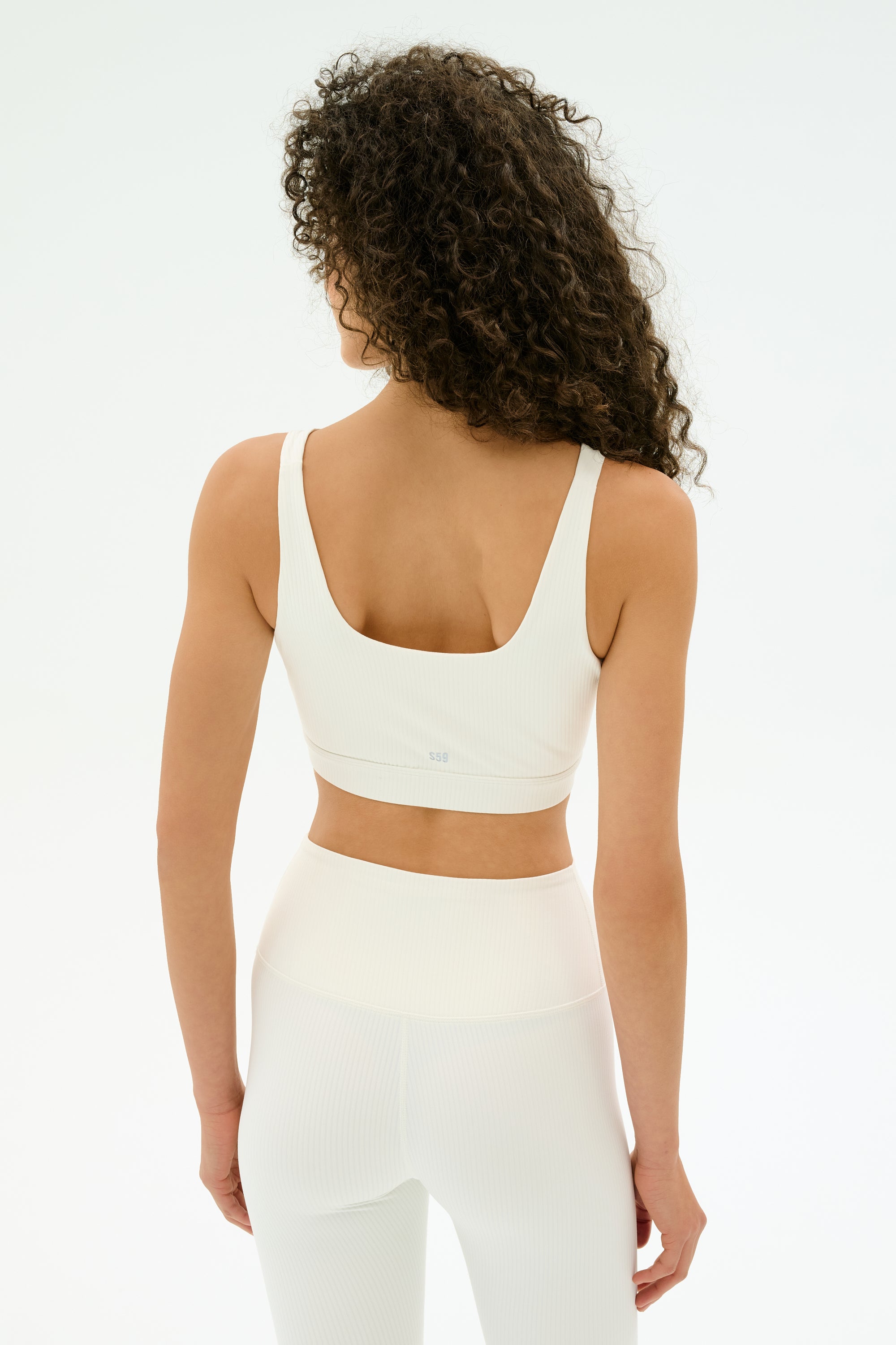 A woman with curly hair wears the SPLITS59 Airweight Rib Bra in white, paired with matching high-waisted leggings, standing against a plain white background.