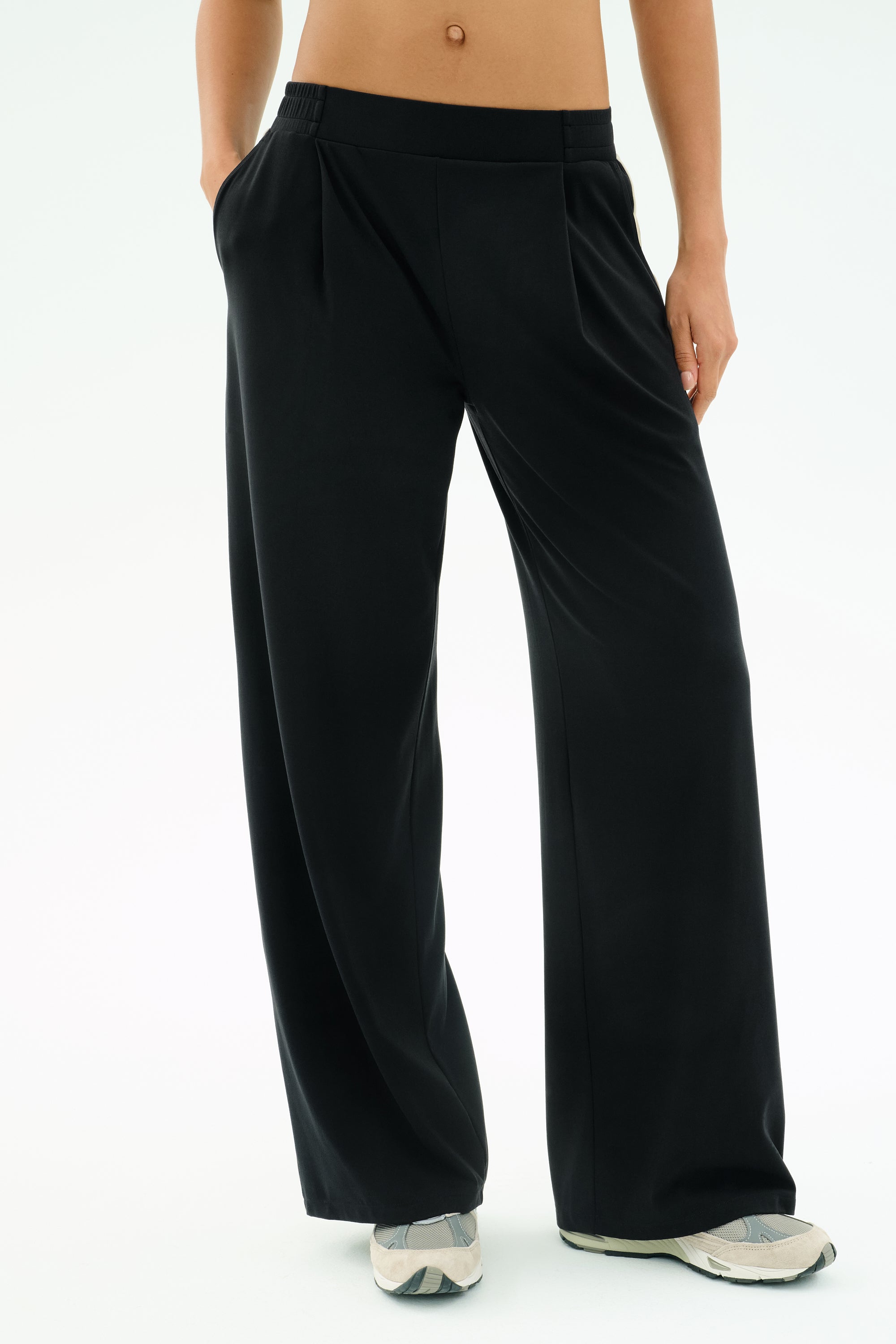 Wearing SPLITS59's Luca Airweight Trouser with Stripe - Black/White and white sneakers, standing with hands in pockets against a plain light background—crafted from ultra-luxe Airweight fabric for all-day comfort.