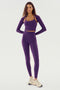 A woman stands against a white background wearing the SPLITS59 Shiri Airweight Shrug - Black Currant, a long-sleeve crop top paired with matching high-waisted leggings. Both pieces are crafted from breathable nylon and spandex fabric, complemented by white and beige sneakers.