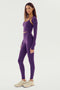 A woman stands in profile wearing the Shiri Airweight Shrug - Black Currant by SPLITS59, paired with matching spandex leggings and white athletic shoes.