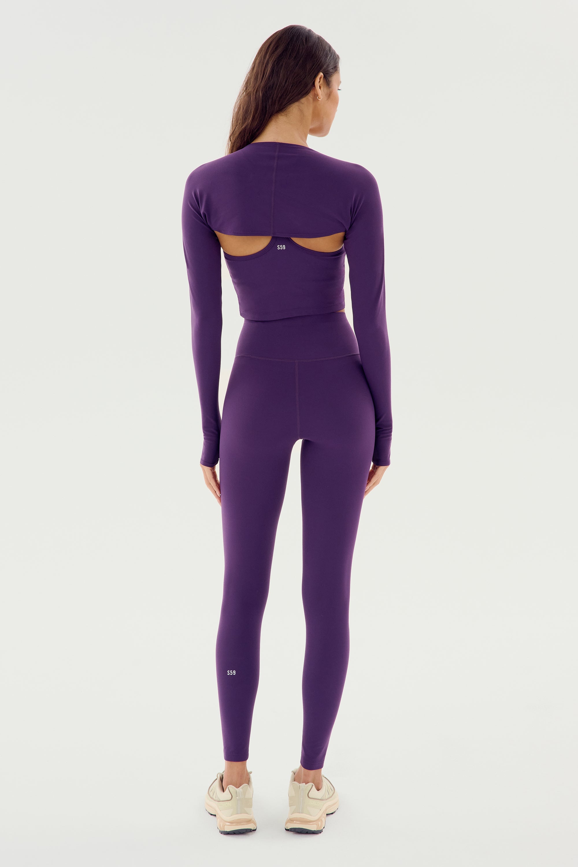 A person with long hair stands facing away, wearing the Shiri Airweight Shrug in Black Currant by SPLITS59 and matching purple spandex leggings paired with beige athletic shoes.