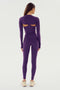 A person with long hair stands facing away, wearing the Shiri Airweight Shrug in Black Currant by SPLITS59 and matching purple spandex leggings paired with beige athletic shoes.