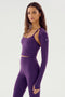 A woman with long hair wears the Shiri Airweight Shrug in Black Currant by SPLITS59, which includes a crop top and matching high-waisted leggings made of a nylon-spandex blend. She poses against a plain background.