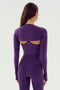 A woman wears the Shiri Airweight Shrug in Black Currant by SPLITS59, featuring a long-sleeve design and constructed from a blend of nylon and spandex, viewed from the back showing the cut-out design on the upper back.
