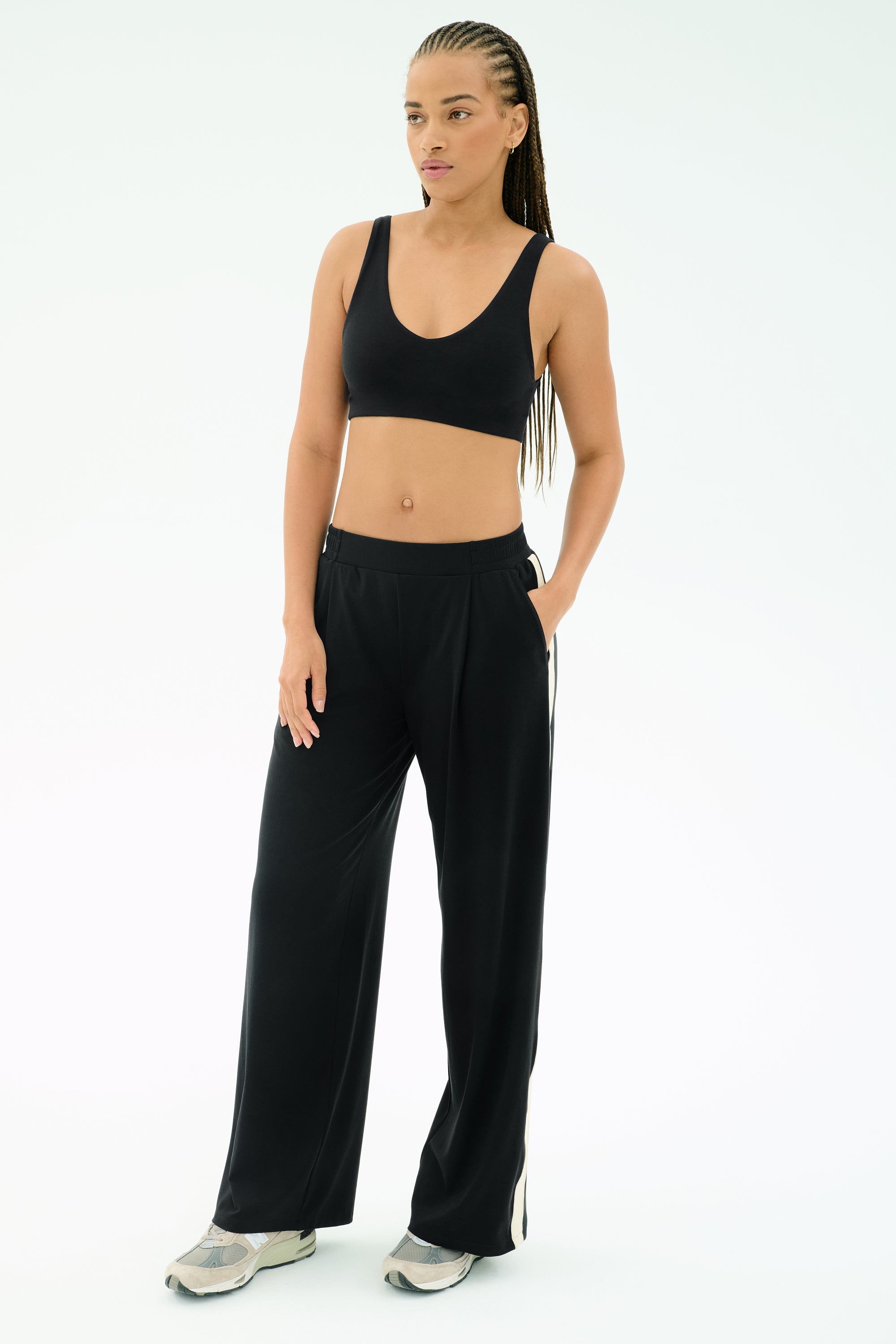 A woman poses against a plain background wearing the SPLITS59 Airweight Scoop Back Bra in black, paired with black wide-leg pants and sneakers, one hand in her pocket.