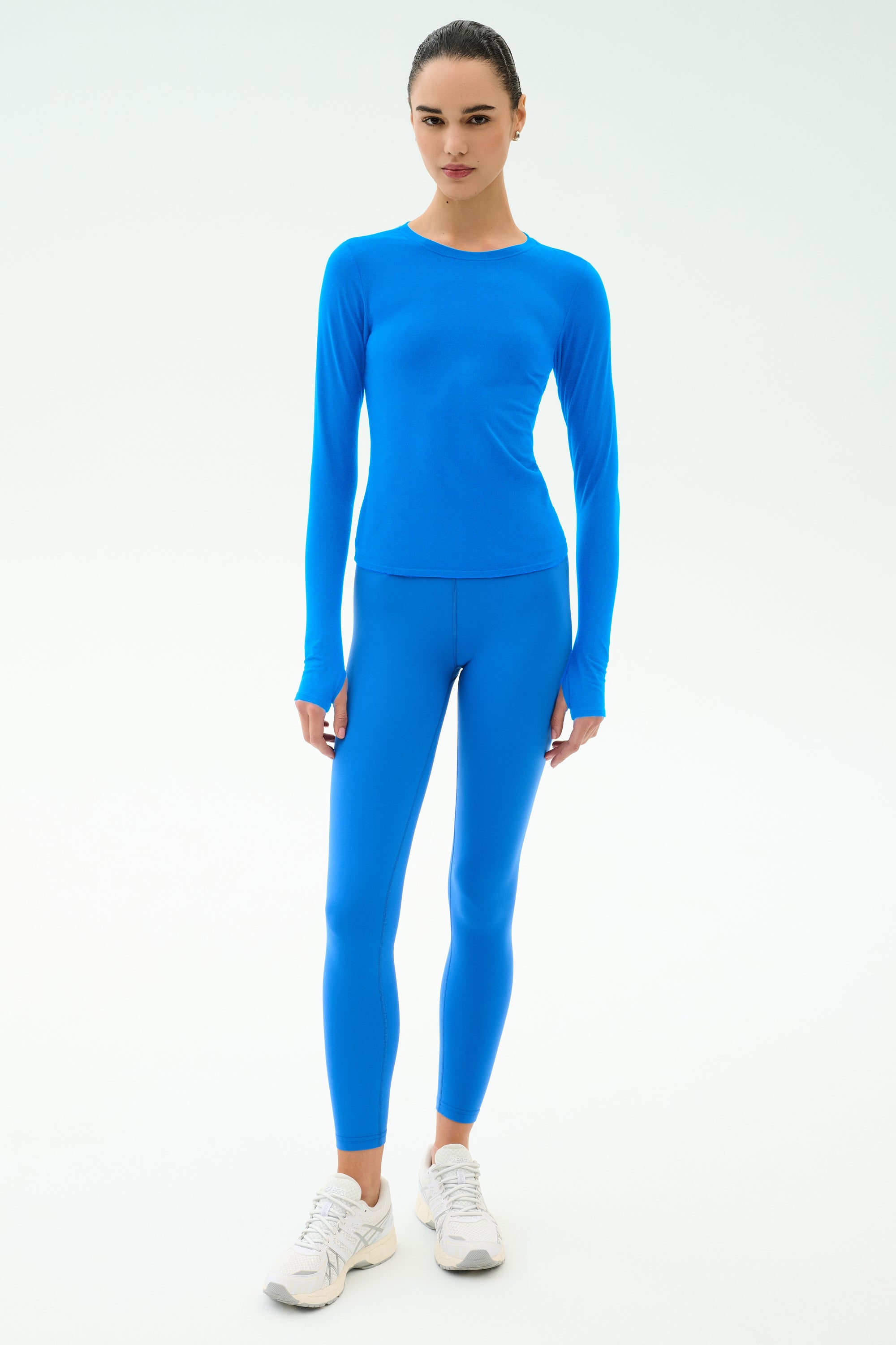 A person stands against a plain background wearing the SPLITS59 Airweight Lite Long Sleeve Waist Length top in Classic Blue, paired with matching blue leggings and white sneakers.