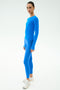 A person stands slightly sideways against a plain white background wearing SPLITS59 Airweight Lite Long Sleeve Waist Length in Classic Blue with matching leggings and white athletic shoes.