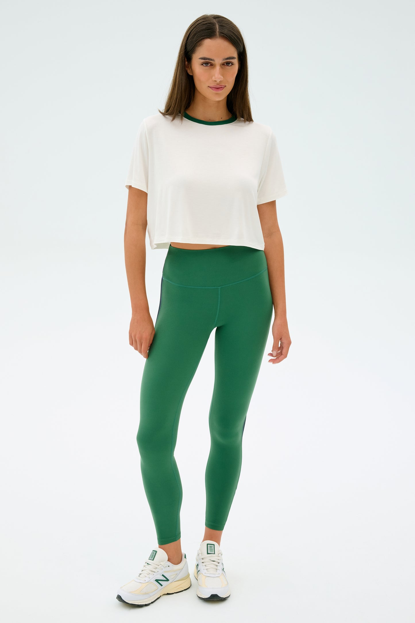 A person stands wearing the SPLITS59 Djuna Crop Ringer in White/Arugula, green elastane leggings, and sneakers against a plain background.