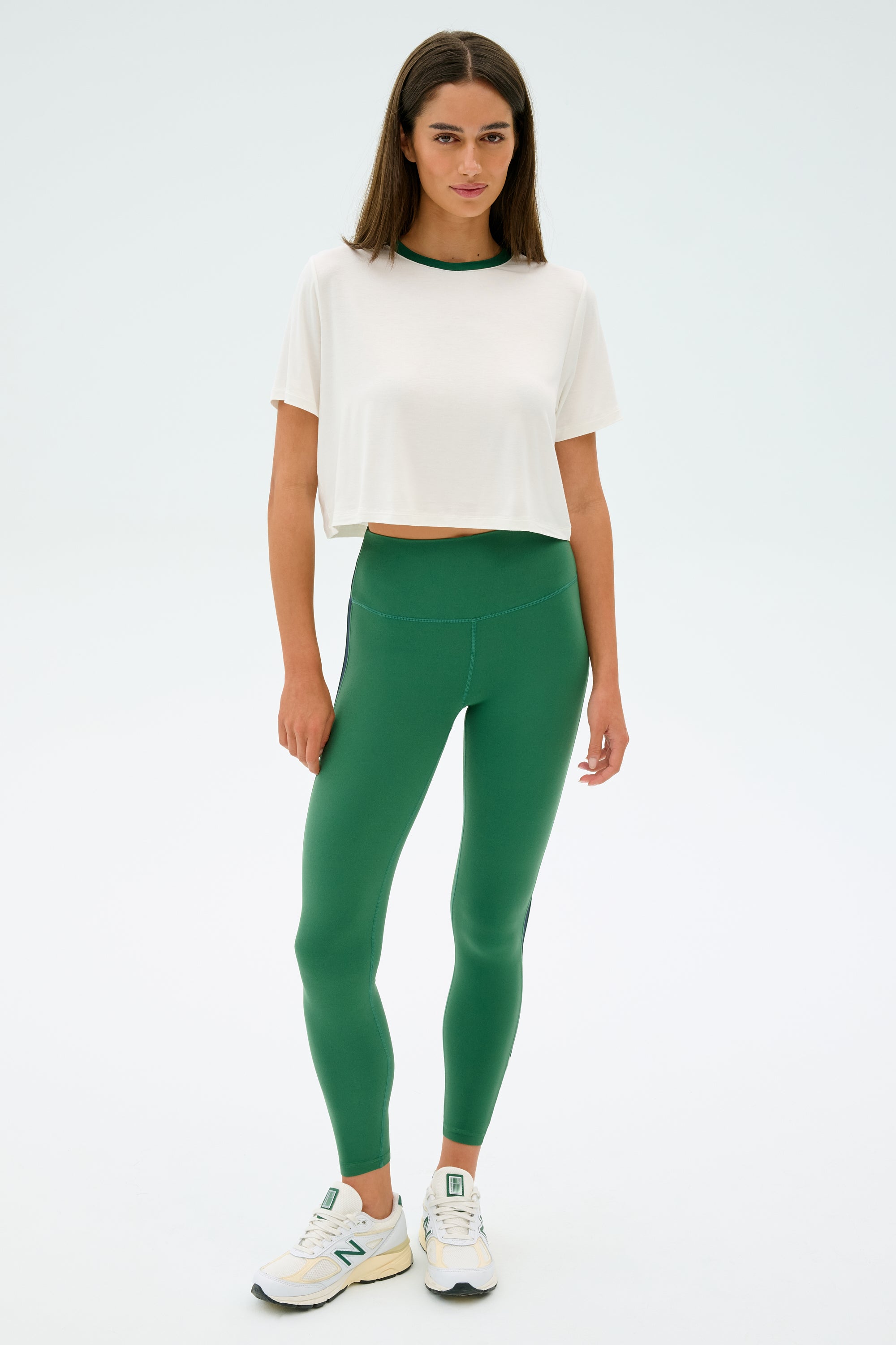 A person stands wearing the SPLITS59 Djuna Crop Ringer in White/Arugula, green elastane leggings, and sneakers against a plain background.