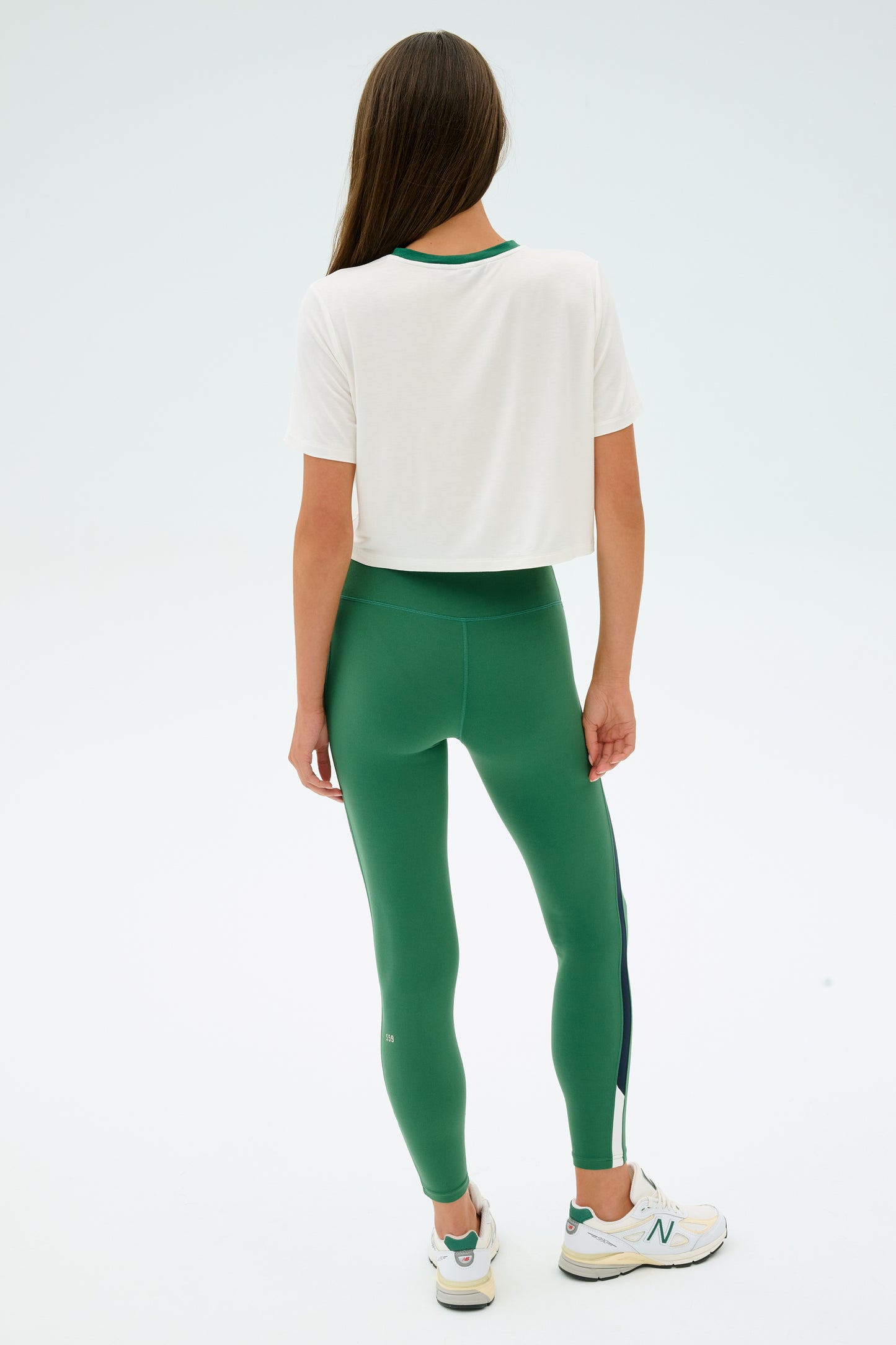 A person with long brown hair, wearing the SPLITS59 Djuna Crop Ringer in White/Arugula and green elastane leggings, paired with white sneakers, stands facing away against a plain background.