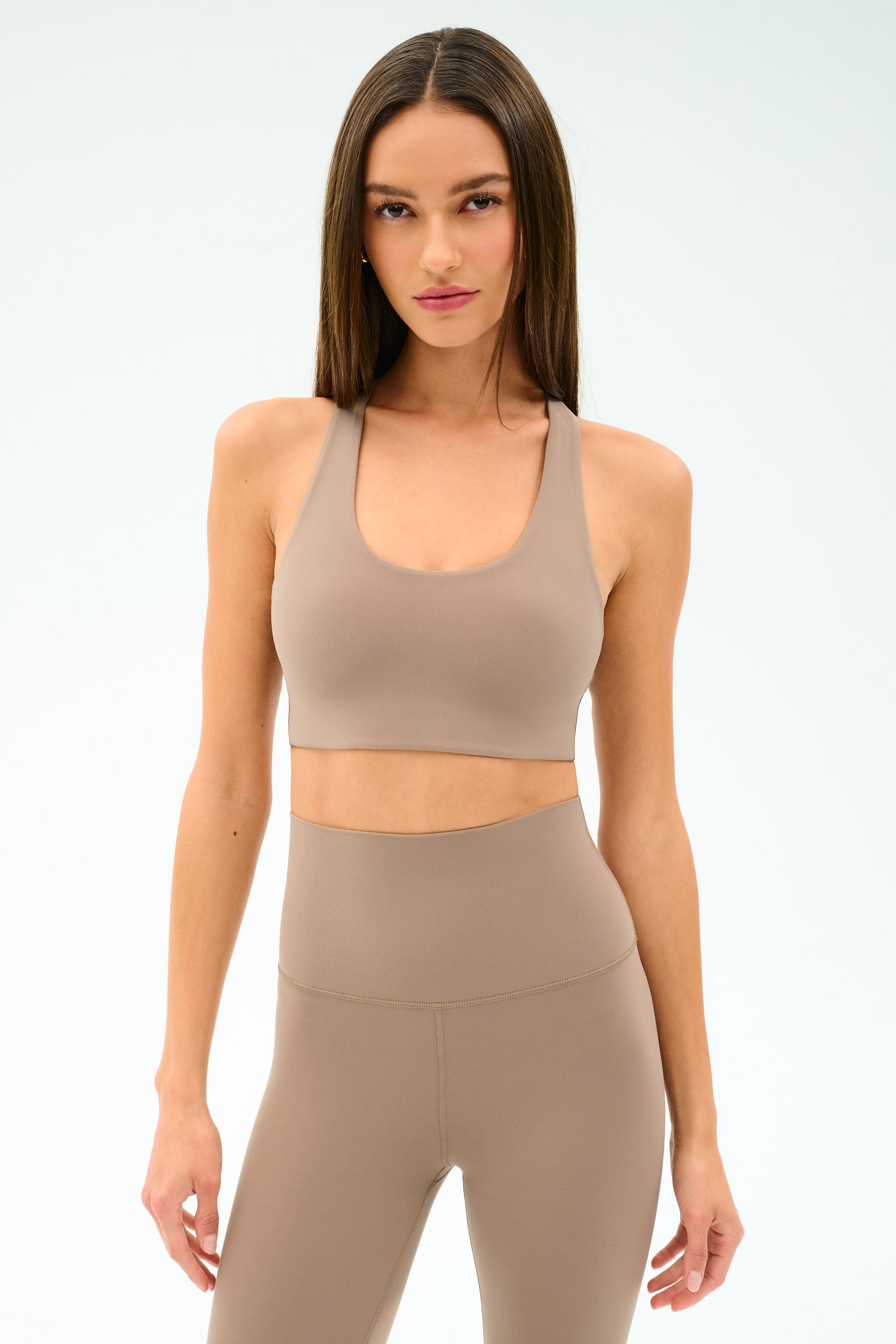 A woman models the SPLITS59 Sara Airweight Bra in Latte with matching high-waisted leggings against a plain white background—ideal for hot yoga or Pilates.