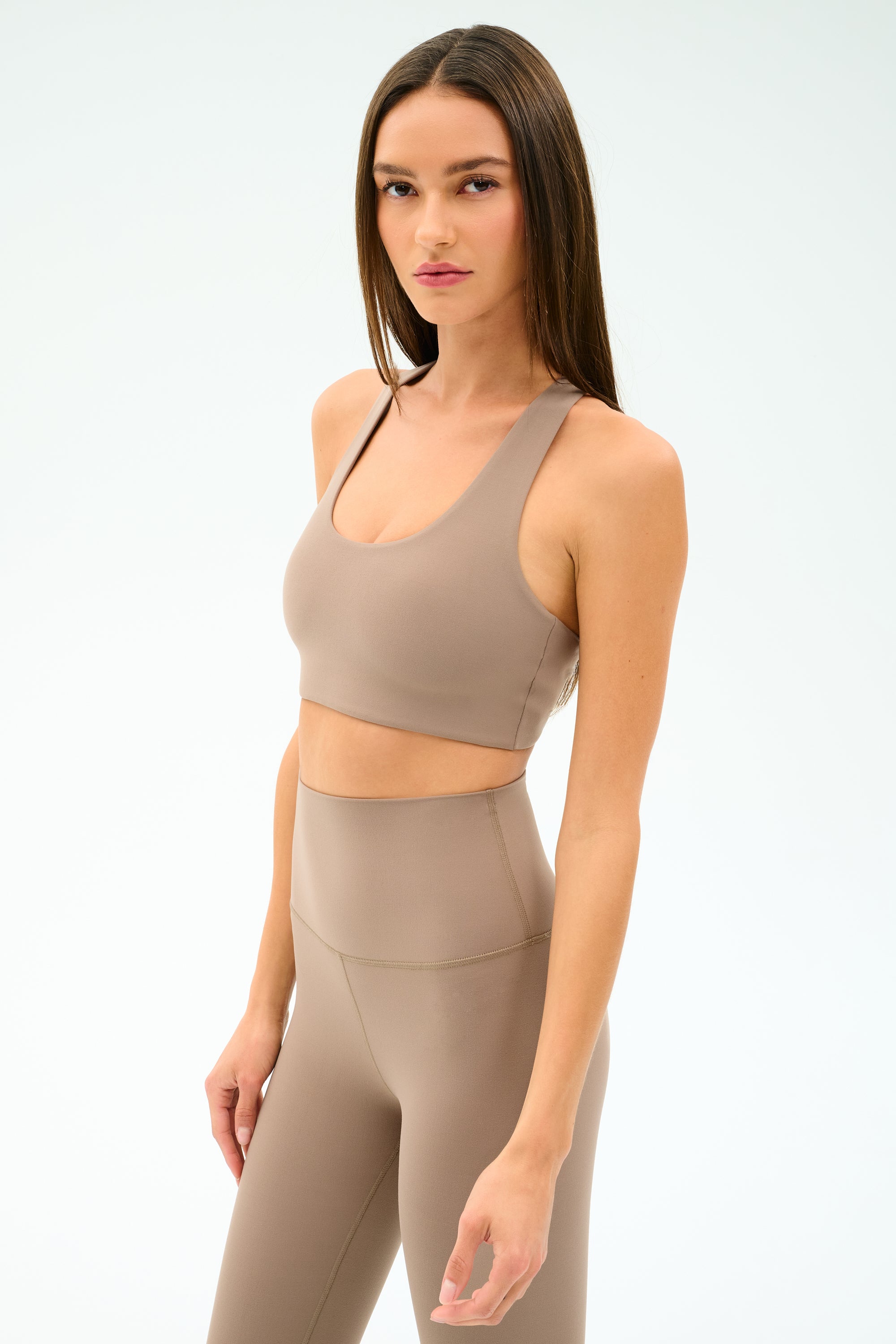 A woman wears the SPLITS59 Sara Airweight Bra in Latte with high-waisted leggings, standing against a plain light background and looking to the side—ideal activewear for hot yoga or Pilates.