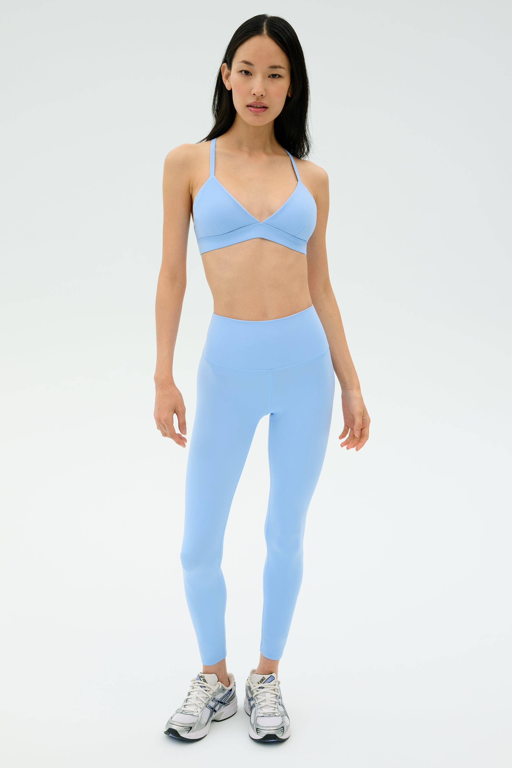 A woman stands against a plain light background, wearing SPLITS59's Sprint High Waist Rigor 7/8 leggings in Skylight and a matching sports bra, paired with silver athletic shoes.