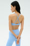A woman wearing the SPLITS59 Theo Rigor Bra in Skylight, featuring crisscross straps, and matching leggings stands with her hands behind her back, facing away.