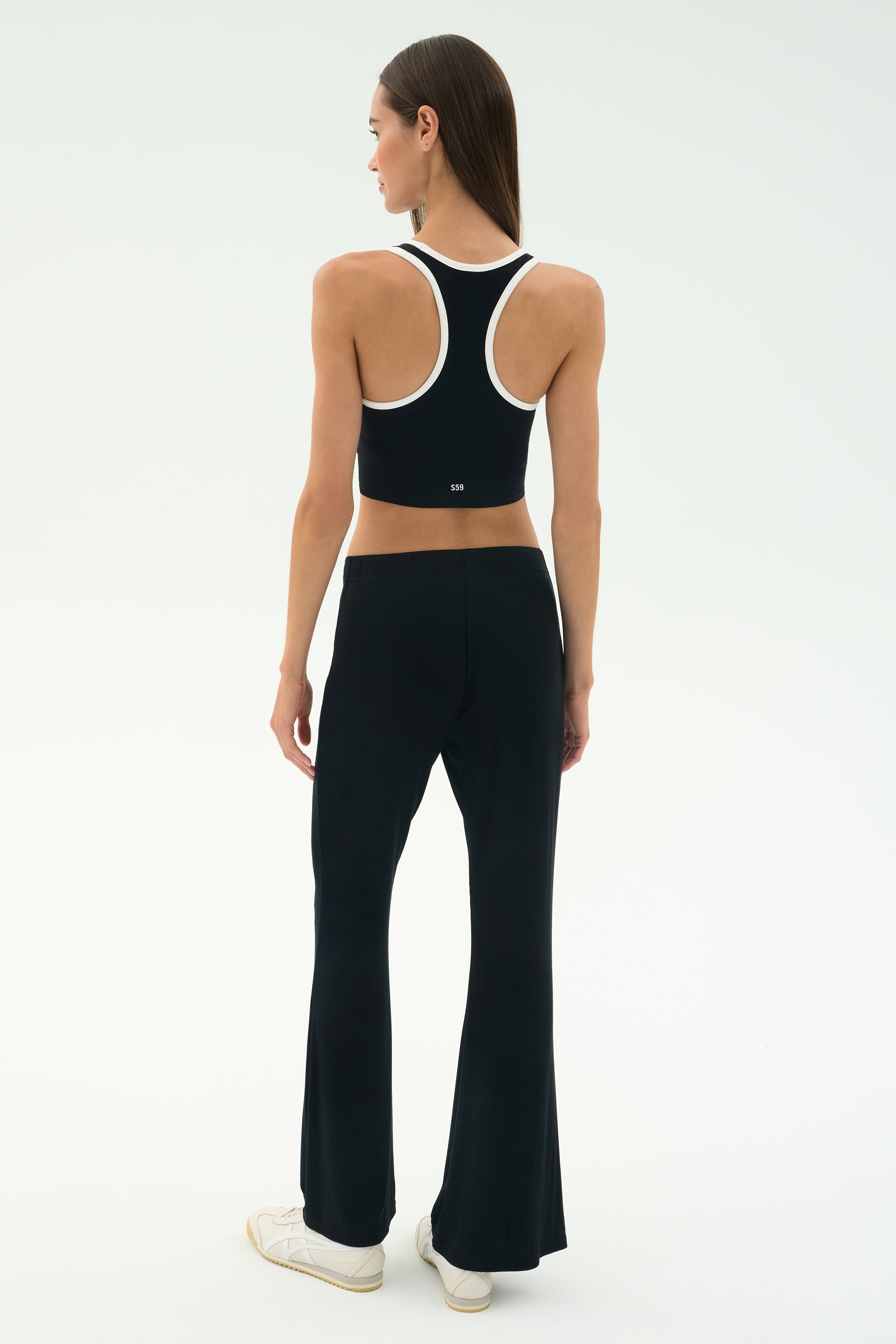 A woman faces away, wearing SPLITS59's Blaire Rib Pant in black with a black racerback crop top, styled with white sneakers against a plain light background.