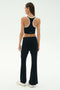 A woman faces away, wearing SPLITS59's Blaire Rib Pant in black with a black racerback crop top, styled with white sneakers against a plain light background.