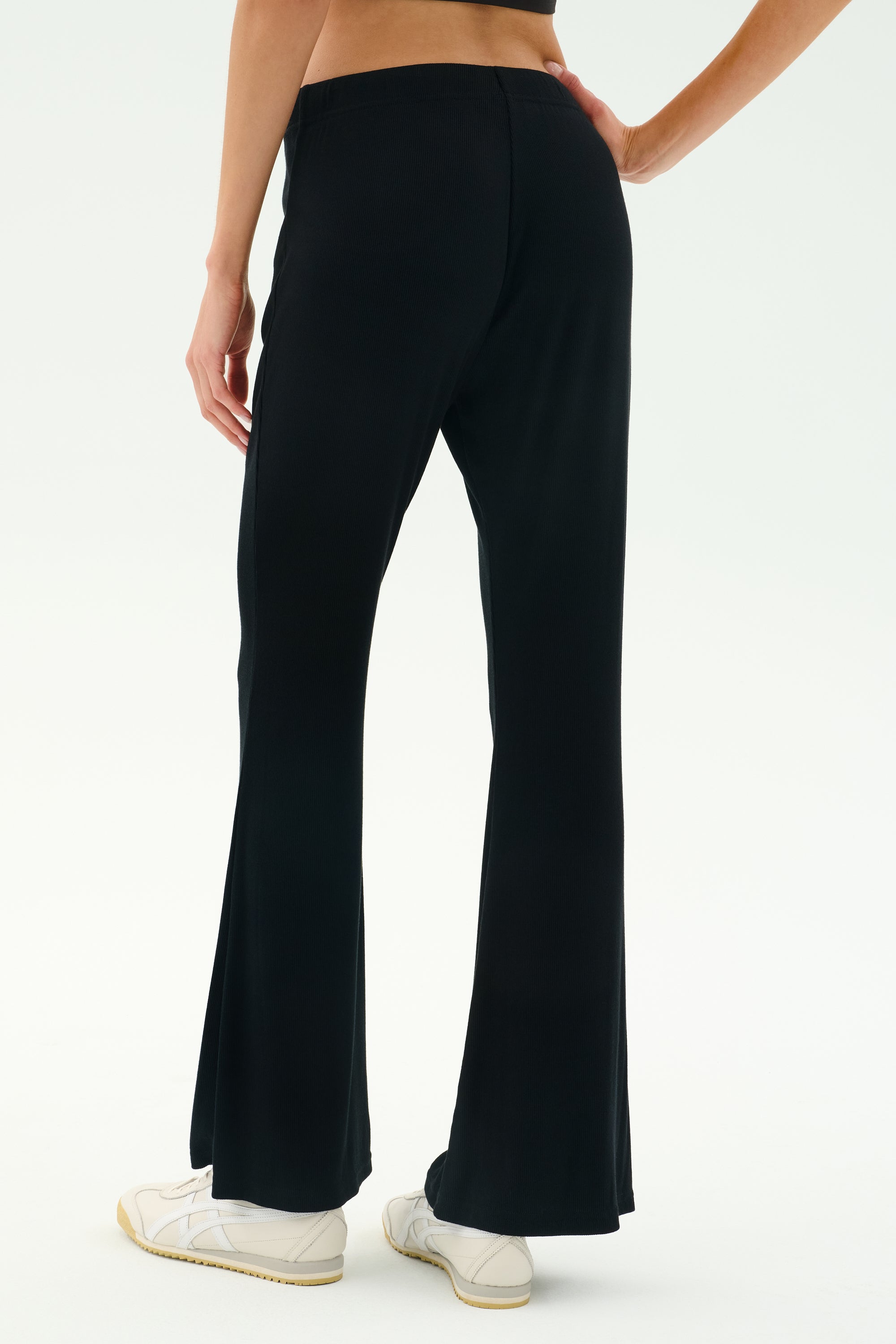 A person wearing SPLITS59 Blaire Rib Pant in black, made with soft modal and spandex, paired with white sneakers, stands on a white background, shown from the back waist down.