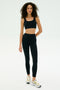 A woman stands against a plain background wearing the SPLITS59 Airweight Rib High Waist 7/8 leggings in black, paired with a matching black sports bra and white sneakers.