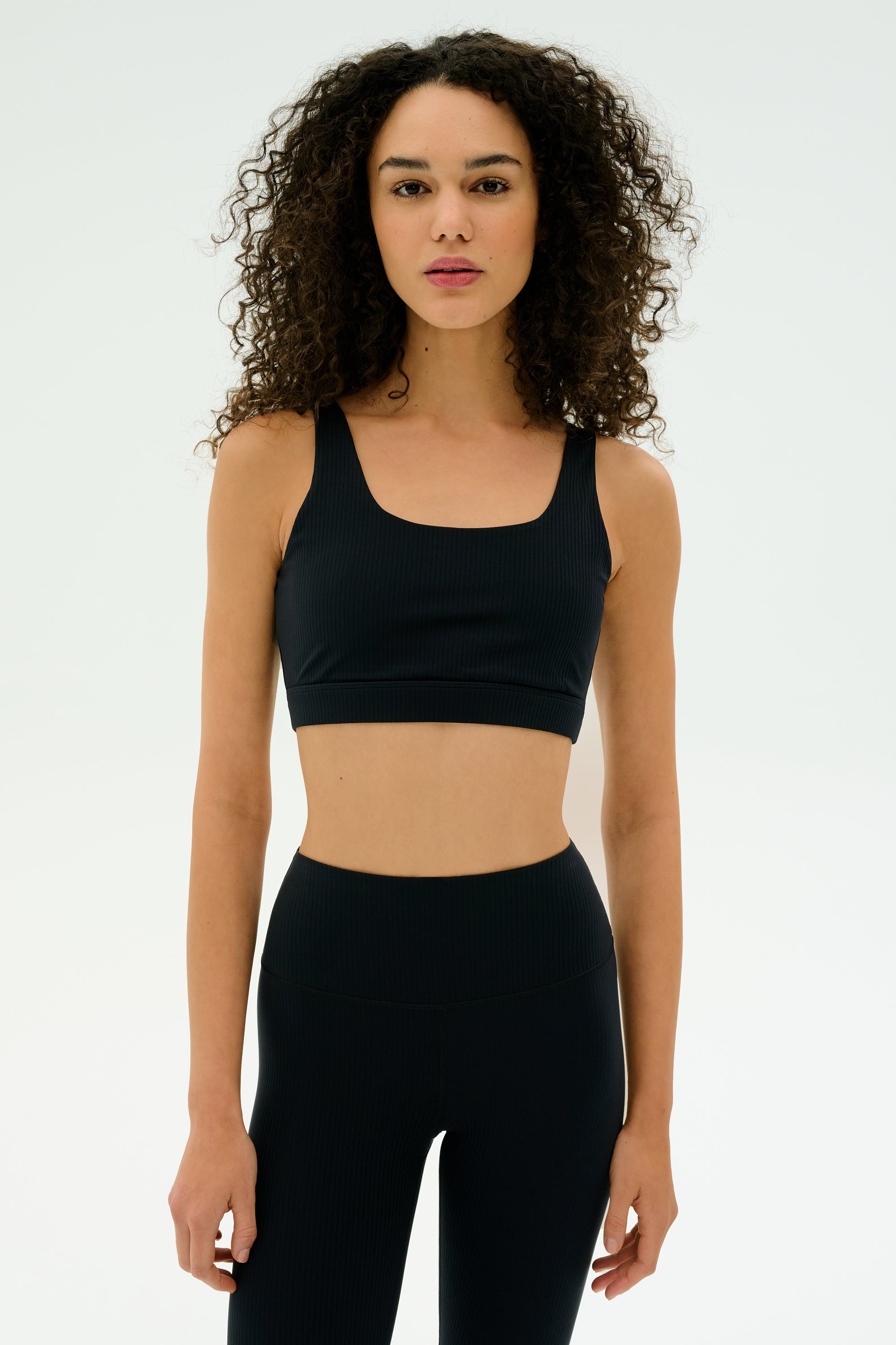 A woman with curly hair wears the SPLITS59 Airweight Rib Bra in black, paired with matching high-waisted leggings, against a plain light background.