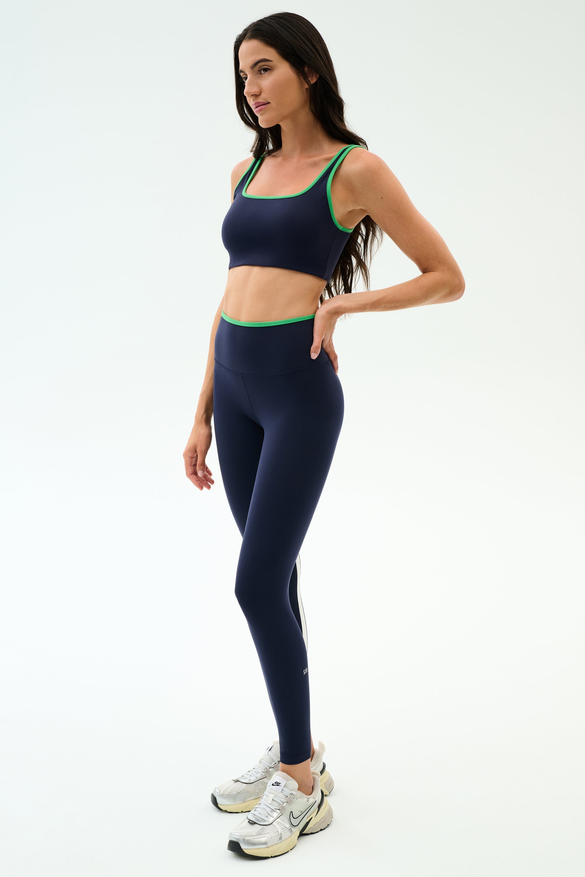 A woman models SPLITS59’s Easton Rigor High Waist 7/8 leggings in Indigo/Grass with a matching sports bra and white sneakers, her long dark hair flowing, set against a plain white background.