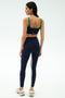 A woman stands facing away, wearing SPLITS59 Easton Rigor High Waist 7/8 leggings in Indigo/Grass with a matching sports bra featuring green trim, a white stripe, and white athletic shoes against a plain backdrop.