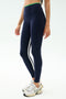 A person wears SPLITS59 Easton Rigor High Waist 7/8 leggings in Indigo/Grass—navy blue with side stripes, made from compressive fabric—with white sneakers, shown from shoulders down against a plain background.