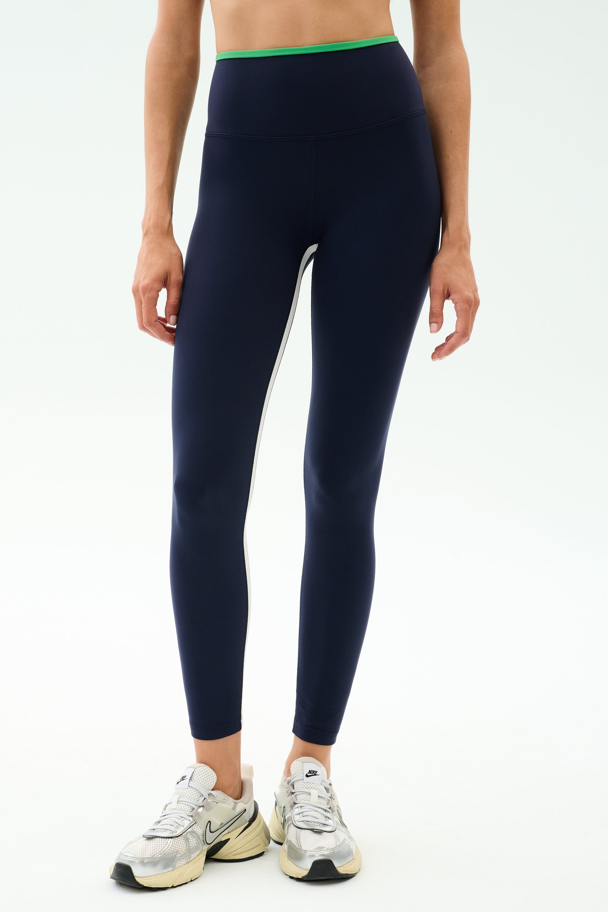 A person wears SPLITS59 Easton Rigor High Waist 7/8 leggings in Indigo/Grass with a green waistband and white side stripe, paired with white Nike sneakers, standing against a plain background.