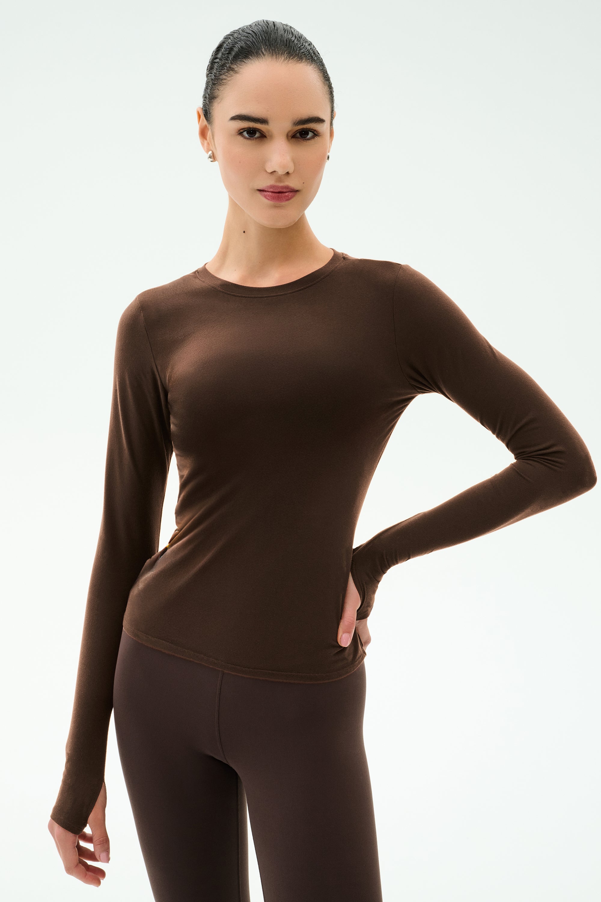 A woman stands against a plain background wearing the SPLITS59 Airweight Lite Long Sleeve Waist Length top in Dark Chocolate, paired with matching leggings, with one hand on her hip.
