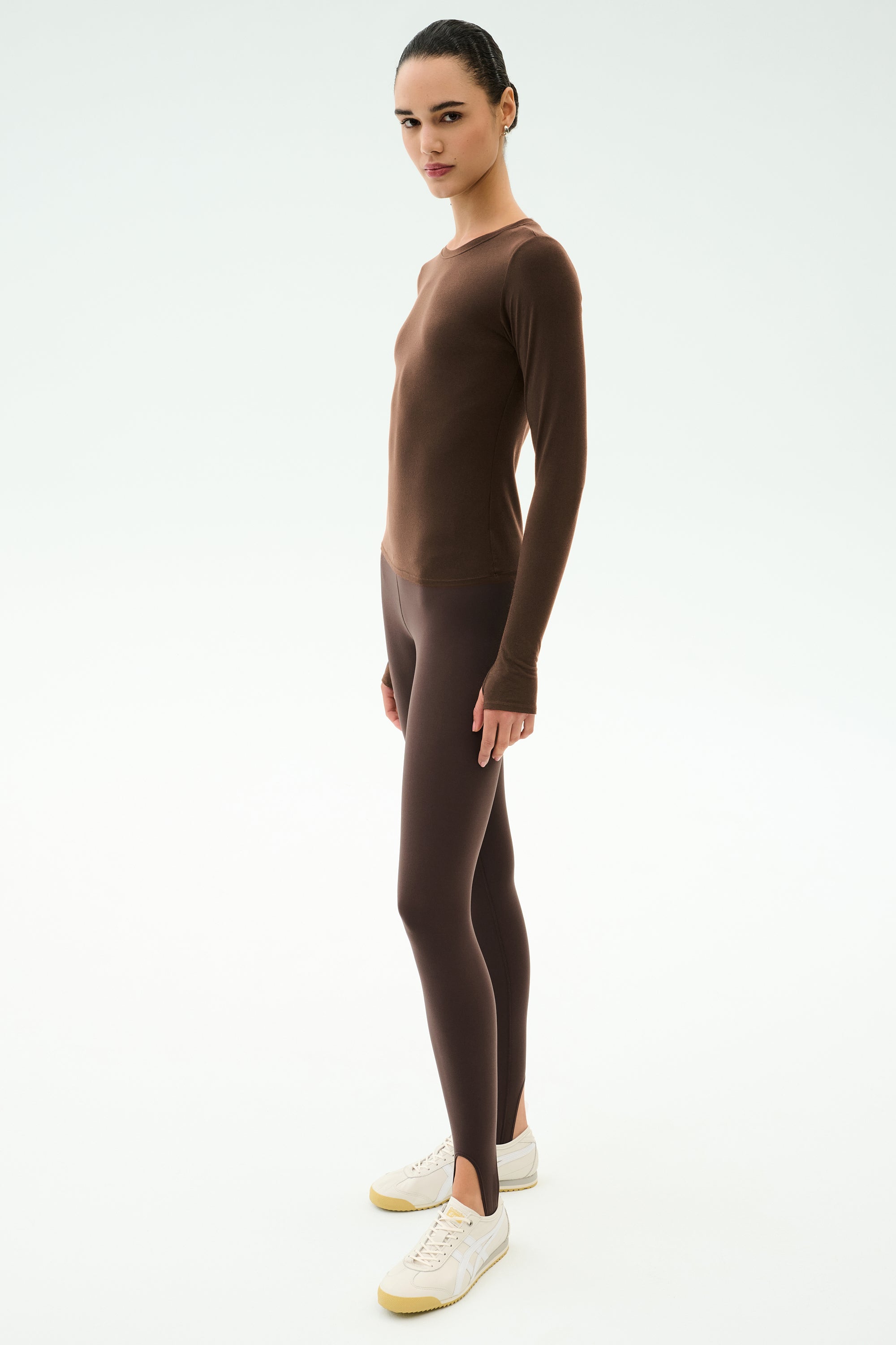 A person stands on a white background wearing the SPLITS59 Airweight Lite Long Sleeve Waist Length top in Dark Chocolate, matching brown leggings with stirrups, and white sneakers.