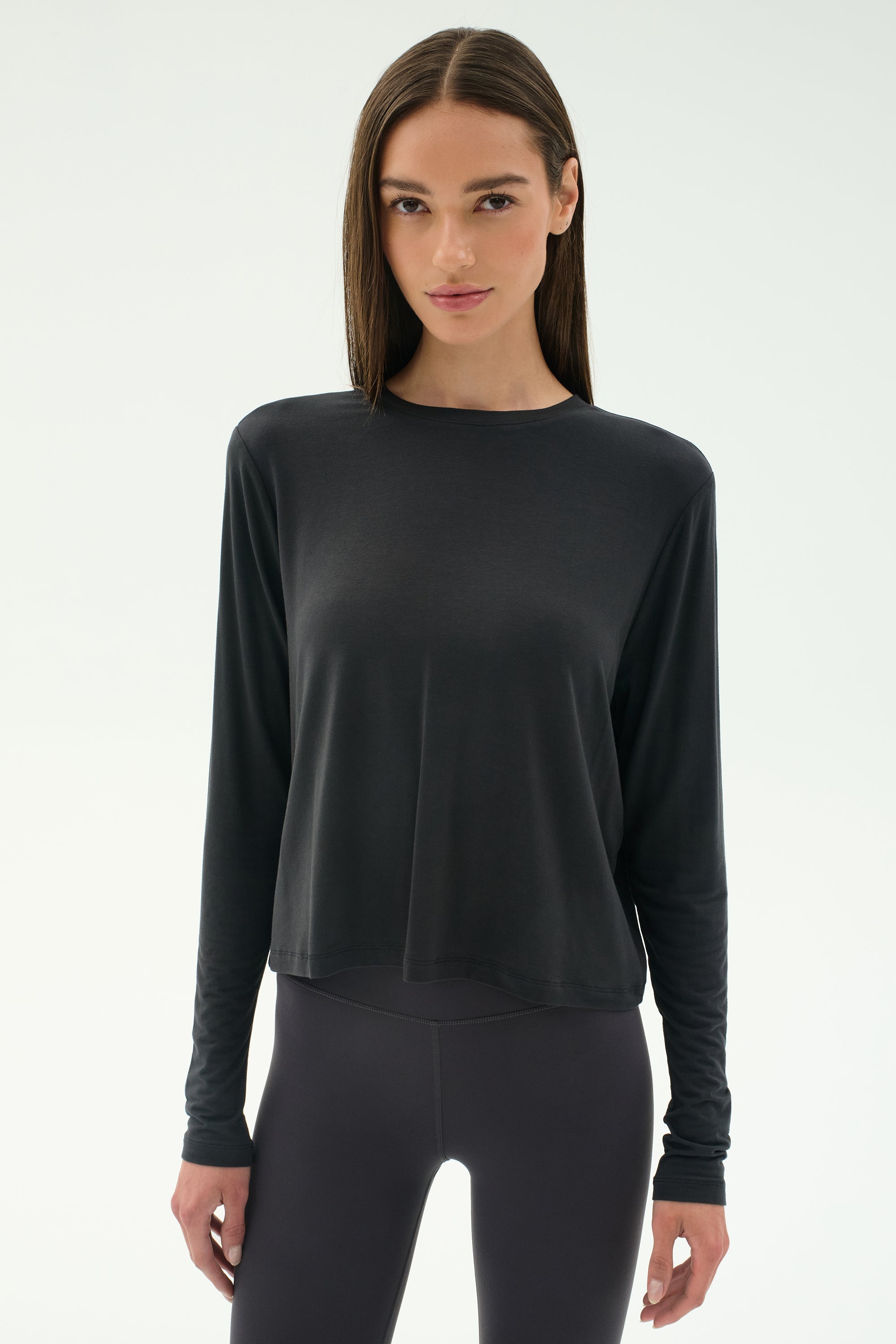 A woman stands facing forward in the SPLITS59 Djuna Long Sleeve - Graphite top and black leggings, set against a plain light background.