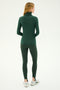 A person with long hair, wearing a supremely soft Jackson Rib Full-Length Turtleneck in Military/White from SPLITS59 and green leggings, is standing and looking to the right. The background is plain and light-colored.