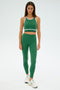 A person wearing the SPLITS59 Layla Airweight Bra in Arugula/Indigo, with matching leggings and white sneakers, stands against a plain background.