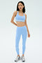 A woman stands against a plain background, wearing the SPLITS59 Miles High Waist Rigor 7/8 leggings in Skylight/White with a matching light blue sports bra and gray running shoes.