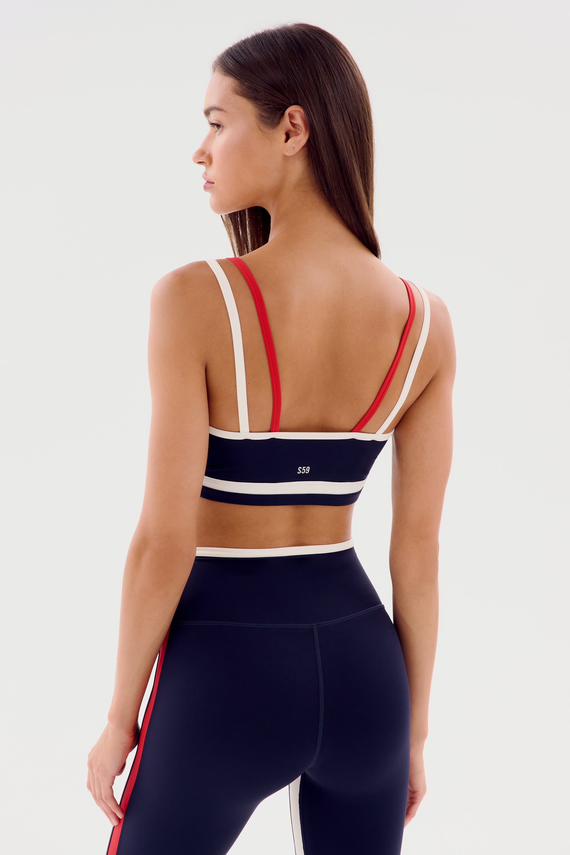 A woman is standing with her back to the camera, showcasing the SPLITS59 Eva Rigor Double Strap Bra - Indigo/White, paired perfectly with matching black leggings featuring red and white stripes.