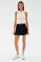 A woman faces forward in a white sleeveless knotted top and the SPLITS59 Venus High Waist Rigor Skort - Black, paired with white socks and sneakers, standing against a plain light background.