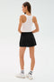 A woman faces away, wearing the SPLITS59 Venus High Waist Rigor Skort in black, paired with a white tank top, white socks, and white sneakers against a plain light background.