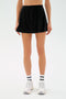 A person wearing the SPLITS59 Venus High Waist Rigor Skort in black, paired with white socks featuring black stripes and white sneakers, stands against a plain light background.