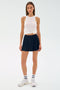A woman faces forward in a white sleeveless top and the SPLITS59 Venus High Waist Rigor Skort in Indigo, paired with white socks and sneakers, set against a plain light background.