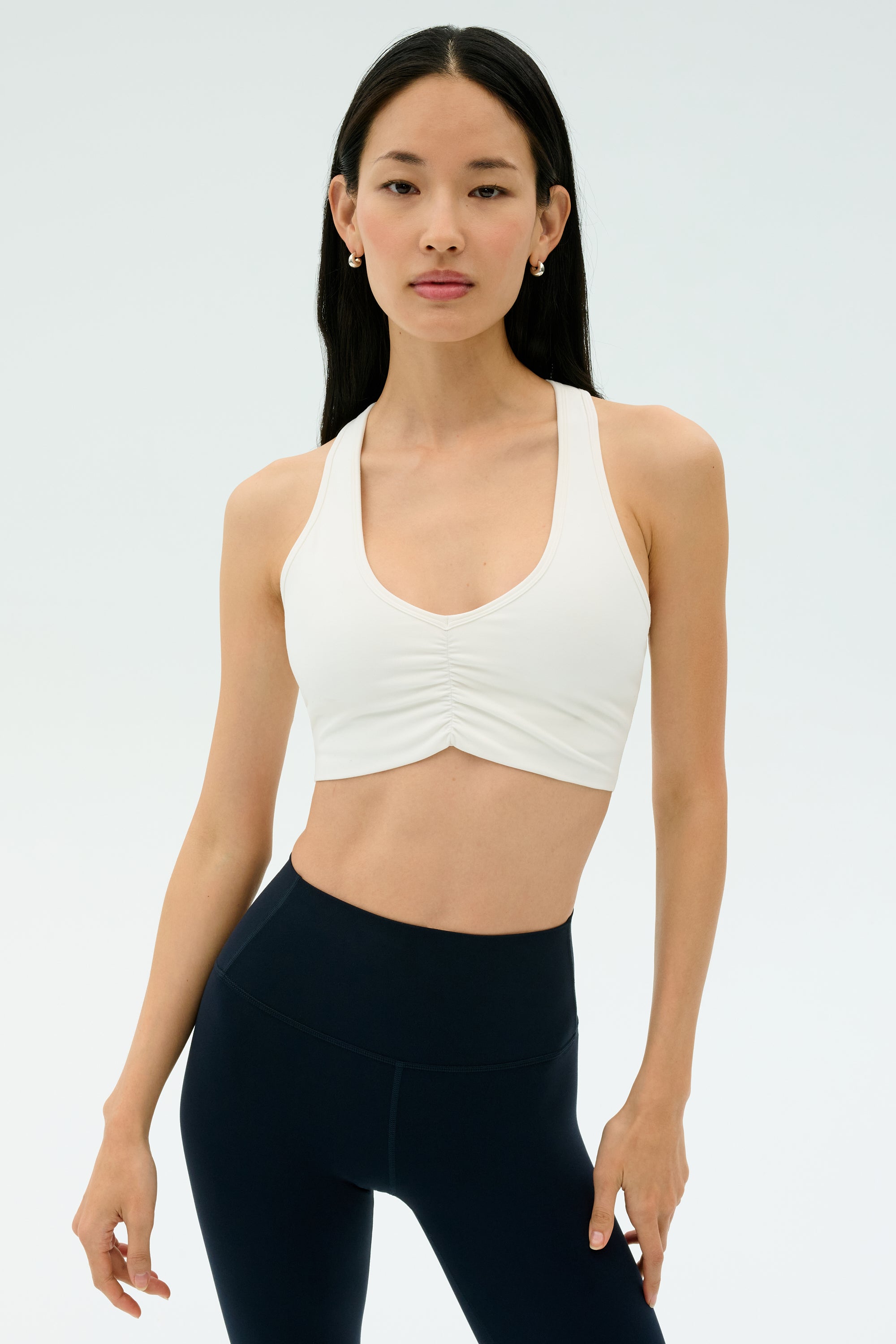 A woman faces forward against a plain light background, wearing the SPLITS59 Airweight Bra with Ruching in white and high-waisted black leggings.