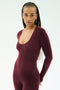 A person with long braids wears the SPLITS59 Chloe Airweight Jumpsuit in Dark Cherry, a fitted long-sleeve piece, standing against a plain light background.
