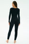 A person with braided hair, barefoot, wears the SPLITS59 Chloe Airweight Jumpsuit in black and stands facing away against a plain white background.