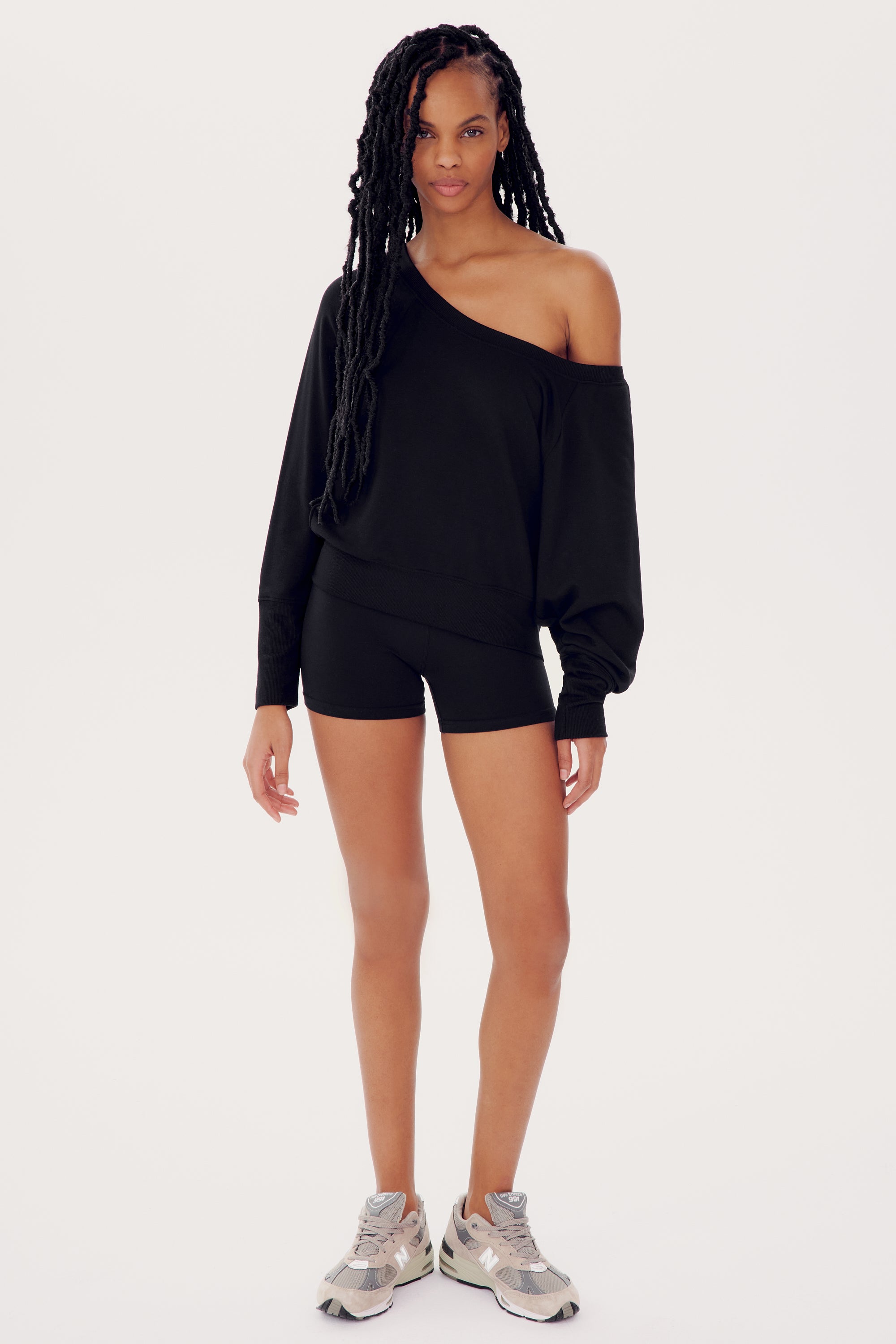 A woman wearing a black SPLITS59 Indy Dolman Fleece Sweatshirt, black shorts, and sneakers stands facing the camera.