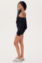 A woman in a black SPLITS59 Indy Dolman fleece sweatshirt and shorts stands profiled against a white background, wearing sneakers.