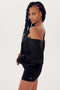 Woman in a black SPLITS59 Indy Dolman sleeve fleece sweatshirt and shorts, looking over her shoulder with a slight smile. Her hair is styled in long braids.