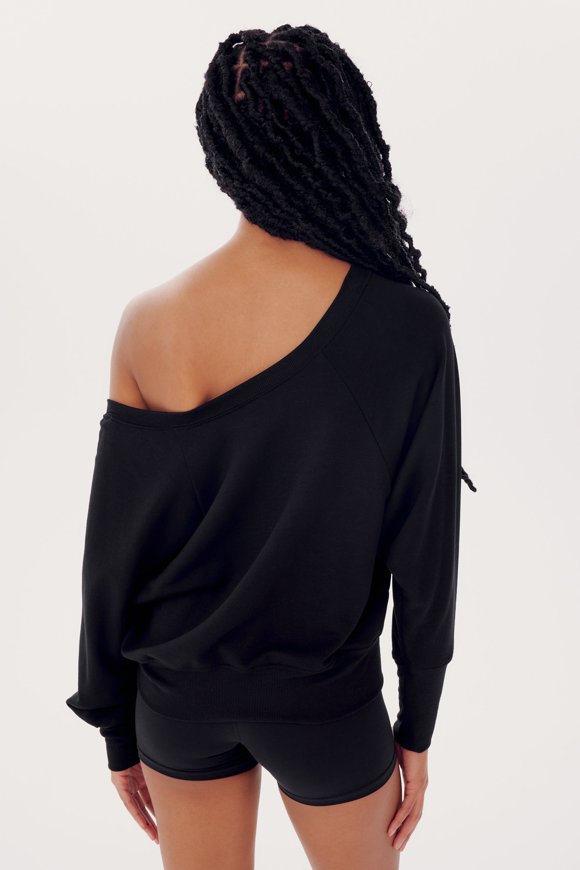 A woman with braided hair sporting a black off-the-shoulder SPLITS59 Indy Dolman Fleece Sweatshirt and shorts, viewed from the back against a white background.