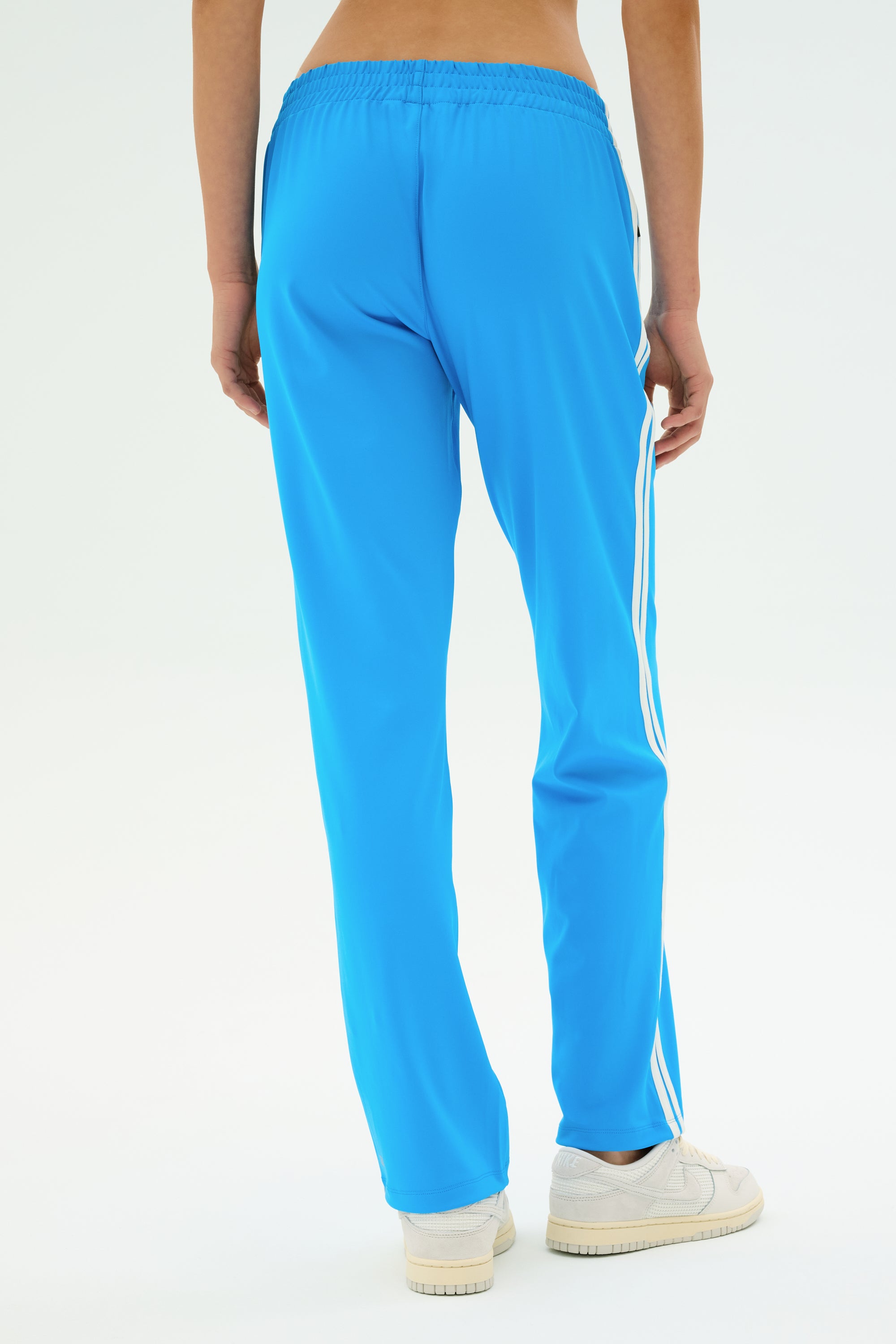 A person is seen from the back wearing SPLITS59 Fox Recycled Techflex Pant in Riviera/White with white side stripes, paired with white sneakers, against a plain light background.
