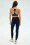 A woman with long brown hair stands wearing SPLITS59's Layla Airweight High Waist 7/8 leggings in Indigo/White and a black sports bra, paired with white sneakers.